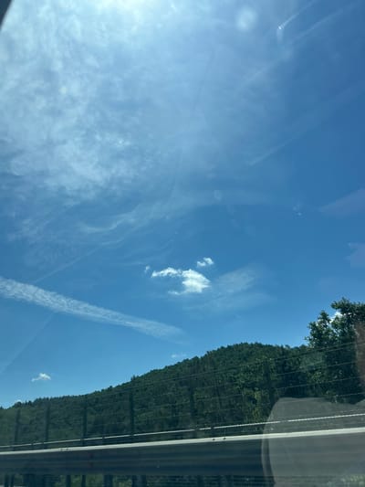 An eye-level, medium shot captures a view of a forested hill under a bright blue sky with scattered white clouds. A metal guardrail and a wire fence line the foreground, obscuring the lower portion of the hill. The scene appears to be taken from a moving vehicle, as suggested by the streaky reflections on the glass surface and the slight blur in the image. The sun is high in the sky, casting a bright light and creating some glare. The location is Novi Han, Bulgaria.