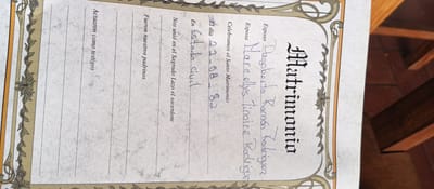 This is a close-up, vertical shot of a marriage certificate. The certificate is ornate, with decorative borders featuring floral motifs and geometric patterns. The background is textured, resembling aged paper.
The primary focus is on the handwritten entries on the certificate, which are in Spanish. The title "Matrimonio" is prominently displayed in a stylized font. Below this, fields are filled in, indicating the names of the groom, "Dagoberto Ramón Rodriguez," and the bride, "Mareelys Tinaire Rodriguez." The date of the marriage is recorded as "21-08-82," and it is noted that the ceremony took place "En Estado civil" (in civil status). Other text on the certificate includes phrases like "Celebramos el Santo Matrimonio" (We celebrate the Holy Matrimony), "Nos unió en el Sagrado Lazo el sacerdote" (The priest united us in Holy Matrimony), and "Fueron nuestros padrinos" (They were our godparents). The final line reads "Actuaron como testigos" (They acted as witnesses).
The image appears to be taken indoors, with wooden furniture visible in the background, suggesting a room within a building. The lighting is somewhat dim, highlighting the parchment texture and the ink of the handwritten entries. The overall impression is of a preserved historical document.