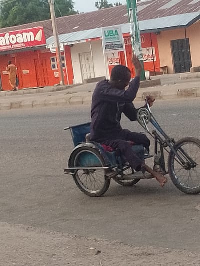 A man is riding a three-wheeled bicycle with a blue cargo area behind him. He is wearing a dark blue garment. The bicycle is on a paved road. In the background, there are buildings with signs. One sign says "afoam". Another sign says "UBA". The overall scene appears to be a street in a town or city. Based on the context of the provided image, this scene is located in Pantami, Nigeria.