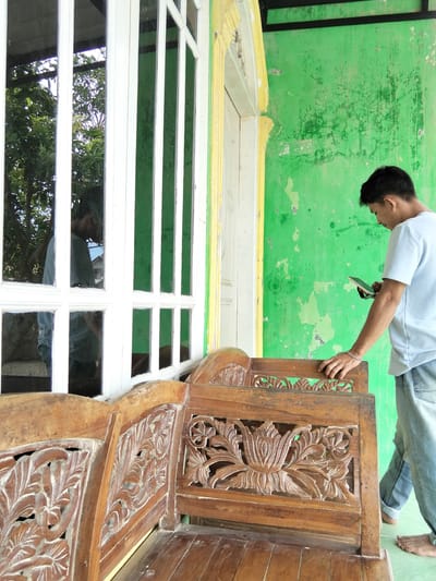 A young man stands near a green wall and a white-framed window in Benuaraja, Indonesia. He is looking down at a dark-colored rectangular object he holds in his right hand. He wears a light blue t-shirt and blue jeans. His left hand rests on the top of a wooden bench with intricate carvings. The bench appears to be aged. The green wall is peeling and stained. In the reflection of the window, another person can be seen standing. The scene is outdoors, with some foliage visible through the window.