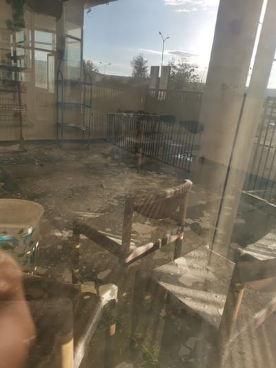 The image captures an interior space, likely a room or enclosed patio, looking out through a large glass window or partition. The scene appears neglected and disused.
**Subjects:**
Indoors, there is a brown wooden chair prominently placed in the foreground, with other less distinct chairs visible. A large, light-colored bucket decorated with blue floral patterns stands to the left. A blue metal shelving unit and part of a red metal rack are also present. The floor is covered with peeling paint or debris.
Through the glass, a sunny outdoor environment is visible, featuring trees, several lampposts, and a metal fence or railing. The bright sunlight suggests a daytime setting.
**Activity or Event:**
No human activity is depicted. The disheveled state of the interior suggests abandonment or a prolonged period of disuse.
**Notable Details:**
The strong sunlight creates reflections on the glass, partially obscuring the view but also highlighting dust and dirt on the surfaces. The overall impression is one of decay and neglect. The outdoor sky is bright with some scattered clouds.
**Visible Text:**
On a sign or poster visible through the glass, near the blue shelving unit, Cyrillic text can be seen, which reads "ГОРНА БАНЯ" (GORNA BANYA).