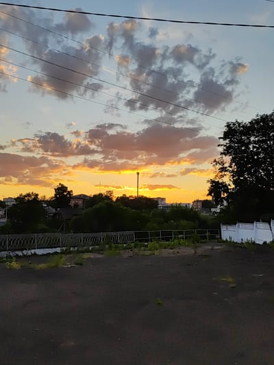 A sunset scene is captured from a low angle, looking towards a horizon line that is obscured by trees and buildings. The sky is dominated by dramatic clouds, illuminated by the setting sun, which casts a warm, orange glow. In the foreground, there is a dark, asphalt-covered area with scattered patches of dry grass and small weeds. A decorative metal fence runs horizontally across the middle ground, separating the paved area from a strip of greenery and vegetation behind it. Beyond the fence, a dense line of trees forms a backdrop, interspersed with glimpses of houses and buildings. A tall, slender communication tower is visible rising above the trees to the right of the center. To the far right, a white, low-rise structure with several sections is partially visible. The overall impression is one of a slightly neglected urban or suburban fringe at dusk. The location in Korosten, Ukraine is not discernible from the visual elements of the image itself.