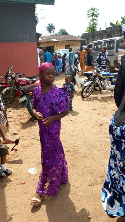 This outdoor photograph, taken in Agburu Uke, Nigeria, depicts a daytime social gathering on unpaved, sandy ground. The central figure in the foreground is a young girl, dressed in a vibrant, floor-length purple gown adorned with sequins and a matching headwrap. She wears light-colored sandals and has a neutral expression, looking to her right. Partially visible to her left are another person's feet in black shoes, and to her right, a person in a blue and white patterned skirt.
In the midground, several motorcycles are parked, including a red Qlink motorcycle behind the girl with a partially visible "ABIA" license plate, and a blue Qlink motorcycle further to the right. A small child in a patterned blue outfit stands behind the girl. Beyond these, a group of adults and children are gathered, some seated on benches, others standing and conversing. Their attire ranges from casual to semi-formal, including traditional wear like an orange outfit on one man.
The background features various vehicles, including a white van with partially visible text "SOLAS AS MOTOR..." and "UMTC". Simple buildings with corrugated iron roofs are visible under a bright, sunny sky. Palm trees and other vegetation dot the landscape, characteristic of a rural or semi-urban African setting. The presence of strong shadows suggests a clear, bright day. The overall atmosphere is one of a calm community gathering.