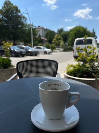 A white coffee cup sits on a dark grey table in an outdoor setting. The cup has the words "your moment. awake." printed in red on the inside rim. In the background, several cars are parked on a street. Trees and buildings are visible, along with a white van. The sky is blue with some clouds. The scene appears to be a street cafe in Strășeni, Moldova.