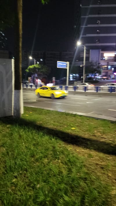 At night in Jiulongpo District, China, a yellow taxi cab travels on a street, passing a guardrail. The background reveals a cityscape illuminated by streetlights. The roadside features a grassy area.