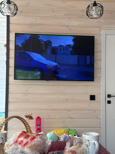 The image shows a modern living room with light-colored wooden paneled walls. A flat-screen television is mounted on the wall, displaying a blue car on a street at dusk. Above the television, two black pendant lights with intricate circular designs hang from the ceiling. To the right of the television is a white door with a black handle and a black light switch.
In the foreground, a table is set with various items. A wicker basket sits on the table, containing a plastic-wrapped food item with red markings. Next to the basket are several stuffed toys, a pink toy, a golden decorative item, and a white mug with a dog illustration. Another white mug is visible to the right, partially obscured by the basket. The overall impression is of a cozy, lived-in space, possibly in a house or apartment in Дачное, Kyrgyzstan.