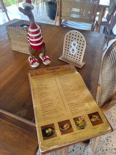 A wooden table in the city of Tegallalang, Indonesia, is set with a menu, a decorative wooden duck figurine wearing a red and white striped shirt and a red hat, and a small woven basket. The menu, which is open, displays various food items. A table number sign with the numeral '8' in white is positioned on the table. The environment suggests an outdoor seating area of a cafe, with glimpses of chairs and plants in the background.
