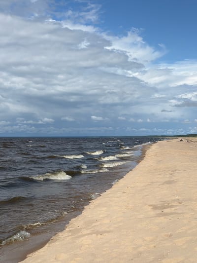 This is a full shot photograph of a beach scene on a cloudy day. The lower left quadrant shows dark, choppy water with white-capped waves rolling towards a sandy shore. The beach occupies the right half of the frame, appearing tan and lightly textured with small scattered white shells. In the distance, along the coastline, a treeline is visible, and a few small figures can be seen sitting or standing on the sand. The upper portion of the image is dominated by a dramatic sky filled with heavy, grey clouds, interspersed with patches of blue sky. The location is identified as Gauja, Latvia.