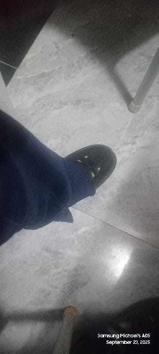 An indoor scene in Uweyoba, Nigeria, features a human foot resting on a light-colored, marble-patterned tiled floor. The foot, positioned in the mid-left quadrant, is clad in dark blue fabric, likely trousers or sweatpants, and a dark-colored, possibly black, slip-on shoe with multiple circular perforations across its upper surface. The floor exhibits a greyish hue with discernible veining and grout lines; minor surface debris is present. Shadows are prominent, suggesting an interior setting with directional lighting. A metallic cylindrical leg, consistent with furniture, is visible vertically in the upper right of the frame, casting a shadow. Another similar metallic leg with a light brown cap is partially visible at the bottom center. A bright, reflective area appears on the floor in the lower right quadrant. A digital timestamp overlay reads "Samsung Michael's A05 September 23, 2025." No other individuals or active interactions are depicted.