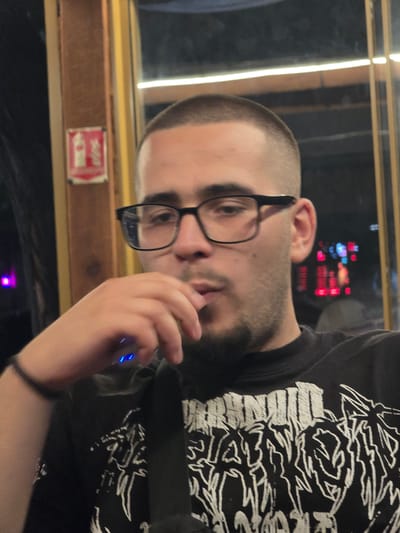 A male individual is depicted in an indoor setting within the city of Lom, Bulgaria. The subject has closely-cropped hair, dark-framed eyeglasses, and facial hair consisting of a beard and mustache. He is wearing a black t-shirt with a white graphic design, which includes partially discernible text and abstract elements. His right hand is positioned near his mouth, holding a white and blue electronic device with two illuminated blue indicator lights. His mouth is slightly open, consistent with inhalation or exhalation from the device. A dark, thin bracelet is visible on his left wrist. The background presents an interior environment with dim, warm lighting and reflective surfaces. To the left of the subject, a red sign with white iconography, consistent with a fire extinguisher symbol, is mounted on a wooden or brown panel. A faint purple light source is also visible in the left background. Through a reflective surface to the right, blurred red and blue lights are discernible. A prominent horizontal light reflection extends across the upper portion of the background.