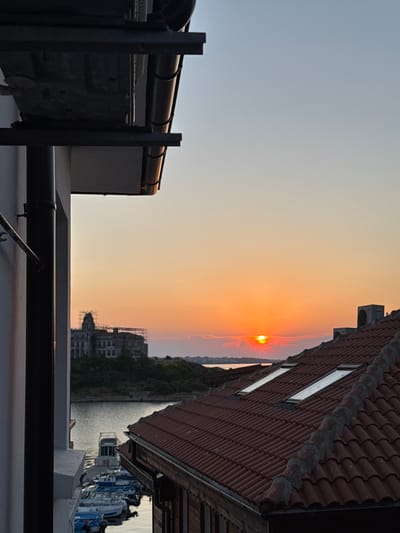 The image depicts a sunset scene in Sozopol, Bulgaria. The sun is low on the horizon, casting a warm orange glow across the sky. The foreground features rooftop elements, including pipes, railings, and part of a building with a tiled roof. To the right, a building with a red-tiled roof and windows is visible. In the background, across a body of water, there is a tree-covered hillside and buildings, some of which appear to be under construction. Boats are docked along the waterfront on the right side of the image. The overall mood is peaceful and serene, characteristic of a sunset. The time of day is clearly evening, as indicated by the setting sun.