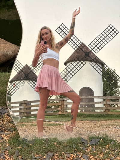 In the city of Thôn 5, Vietnam, a blonde woman with tattoos on her arms and legs is taking a selfie in a cracked mirror. She is wearing a white crop top and a pink pleated skirt. The mirror reflects two white windmills with dark brown roofs and black sails, a wooden fence, and a body of water. The ground around the mirror is covered in gravel, rocks, and grass. The woman is standing on one leg, with her other leg bent and her arm raised. She is holding a pink smartphone in her hand. The lighting suggests it is daytime, possibly late afternoon.