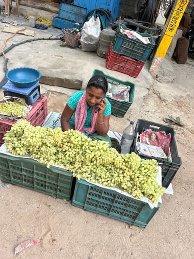 The image depicts a street scene in Puttaparthi, India. An Indian woman is seated on the ground, speaking on a cell phone, while surrounded by crates of green grapes that appear to be for sale. There are at least six crates of grapes in various states of fullness and arranged adjacent to her. A blue scale is near the woman, and newspaper is used to line some of the crates. Other items nearby include water bottles, additional crates, and street debris. The environment appears to be a sidewalk or similar paved area with a dirty surface.
