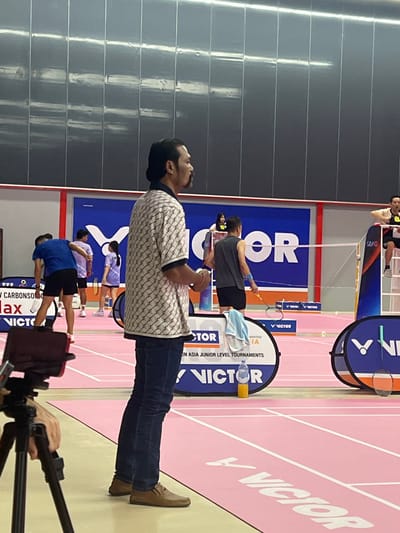 This image depicts a badminton court with several individuals present, likely during a tournament. The court features a pink surface with white lines marking the boundaries. The backdrop includes large blue banners with white text, one of which reads "VICTOR". A prominent VICTOR logo is visible on a smaller circular banner in the foreground.

The central figure in the foreground is a man with dark hair, seen from the side, wearing a patterned short-sleeved shirt and dark blue jeans. He appears to be holding something in his right hand, out of frame. To his left, a camera on a tripod is partially visible, with a hand resting on the tripod.

In the mid-ground, other participants are visible, including individuals in athletic wear on the court. One person, wearing a dark sleeveless shirt and shorts, holds a badminton racket. Another person, a young woman in a purple shirt and skirt, stands on the court. A man in a blue shirt and black shorts is also on the court, facing away from the camera.

Further back, a person is seated on a high chair, possibly an umpire or official, wearing a yellow shirt with the number "8". The environment appears to be an indoor sports facility, with dark, reflective wall panels and overhead lighting. The overall scene suggests a competitive sporting event. The question of location in Ko Samui, Thailand, is not supported by visual evidence within the image itself.