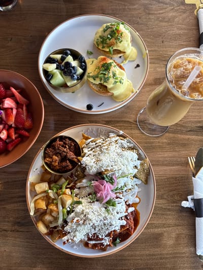 A high-angle shot shows a wooden table set with food, likely in a restaurant in Chula Vista, California, United States of America. On the left, there is a white plate with two servings of what appears to be eggs benedict, accompanied by a small bowl containing blueberries and melon. Next to the plate is a tall glass filled with iced coffee or a similar beverage. To the right, another white plate displays a portion of chilaquiles, a dish of fried tortilla chips in sauce, topped with cheese and possibly other ingredients. A small metal bowl filled with a dark, shredded meat sits next to the chilaquiles, and a bowl of cut strawberries is placed near the bottom of the image. A knife and fork wrapped in a white napkin are placed at the top right. The table exhibits a warm, natural wood grain, and the overall presentation suggests a brunch or lunch setting.