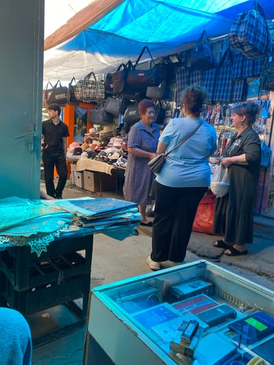 In this street market scene in Gyumri, Armenia, three individuals are the primary subjects. Two women, one in a black dress and the other in a light blue shirt and black pants, are looking at items displayed in a glass case. A third person, a man in a black shirt and pants, is seen from the side, appearing to be in conversation. The market appears to be outdoors, with a blue tarp providing shade. Various objects are visible, including numerous bags hanging from racks, some with checkered patterns and others with the word "SPORT" printed on them. The glass display case contains what appear to be electronic devices, possibly mobile phones, with some bearing labels, one of which reads "SAMSUNG GALAXY A5". There are also stacks of hats and other assorted items for sale. The lighting suggests it is daytime, and the weather seems pleasant. The overall atmosphere appears to be one of commerce and casual interaction within a bustling market environment.