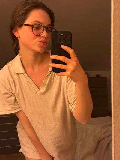 This image depicts a mirror selfie taken indoors in Cava de' Tirreni, Italy. The primary subject is an adult female, positioned from the chest upwards, angled slightly. She wears dark-framed eyeglasses and a light-colored, collared V-neck short-sleeved top. Her lips are pursed in a pouting expression, and a reddish flush is visible on her left cheek. She holds a dark-colored smartphone vertically in her right hand, with its rear camera module and a circular attachment visible. In the background reflection, a dark dresser with multiple drawers is visible to the right, and a portion of a bed with a light-colored duvet or blanket is visible further right. The lighting is warm and dim.