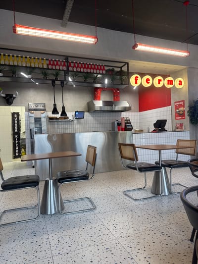 The image depicts the interior of a modern burger restaurant named "ferma" in Kokshetau, Kazakhstan. The establishment features a clean, industrial-chic design with a polished concrete floor, white tiled walls, and stainless steel surfaces. In the foreground, a small wooden table is set with two chairs, each with black leather seats and rattan backs, resting on a chrome base. To the left, a vertical sign reads "BURGER KITCHEN" in white lettering against a black background.
The background reveals a well-equipped kitchen area. A stainless steel counter with overhead shelving displays various condiments, including rows of yellow mustard and red ketchup bottles, interspersed with small potted plants. Above the counter, professional kitchen equipment such as ovens and a range hood are visible. Hanging light fixtures, some industrial black pendant lights and some circular illuminated signs spelling out "ferma," illuminate the space.
On the right side of the image, a menu board displays prices and food items, with a prominent advertisement for a "KOMBO OBED" (combo meal) costing 2990 Tenge. The overall atmosphere suggests a busy, functional, and stylish eatery. No people are clearly visible in the image, though a person may be obscured behind the counter. The scene is brightly lit, suggesting daytime operation.