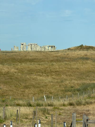 The image depicts Stonehenge, a prehistoric monument located in Wiltshire, England, near West Amesbury. The monument consists of a ring of standing stones, with some stones arranged in horizontal lines atop vertical ones. The stones are weathered and appear to be made of sandstone. They are situated on a grassy hill, which is dry and appears to be covered in tall, golden-brown grass. The sky above is a clear, light blue with some faint, wispy clouds. In the foreground, there is a rustic fence made of wooden posts and barbed wire, with some white and black marker posts scattered throughout. The overall scene is natural and serene, with the ancient monument standing as the focal point against the backdrop of the English countryside. There are no people visible in the image.