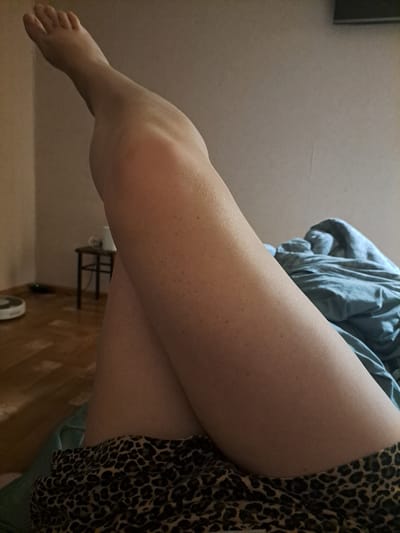 The image is a close-up, low-angle shot of a person's legs, positioned in a bedroom. The person is lying down, with their left leg bent and extended upwards, while the right leg is crossed over it. The person is wearing leopard-print shorts. The background reveals a light-colored wall, a blue comforter, a wooden floor, and a small stool with a white object on it. A white robotic vacuum cleaner is visible on the floor in the distance. The lighting is dim, suggesting it might be evening or a room with limited natural light. The image does not provide any information about the location being in Yessentuki, Russia.