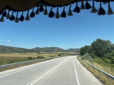 The image shows a view from inside a vehicle, looking out through a windshield that is partially obscured by decorative tassels hanging from the top. The tassels are black and off-white, and appear to be made of yarn or thread.
The scene outside is a rural landscape under a clear blue sky. A paved road curves to the right, with a metal guardrail running along its edge. The road is divided by double white lines. To the left of the road, there is a field of dry grass or crops, and beyond that, rolling hills covered in sparse vegetation and trees. To the right of the road, there are dense green trees and bushes.
The location is identified as Lyra, Greece. The time of day appears to be daytime, likely late morning or early afternoon, given the bright sunlight and clear sky. There are no people or specific actions visible in the image. The environment is natural and appears to be dry and somewhat arid, consistent with a Mediterranean climate.