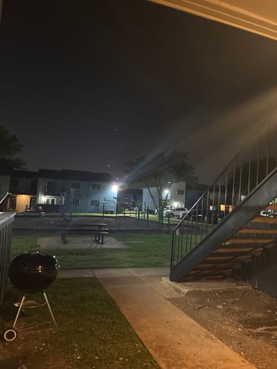 A charcoal grill is positioned on a patch of grass next to a concrete walkway at night. In the background, apartment buildings are visible with some windows lit. A playground area with equipment and picnic tables is situated between the grill and the buildings. An outdoor staircase with a black railing leads up to an apartment on the right side of the frame. The scene appears to be an outdoor common area of an apartment complex in Dallas, Texas.