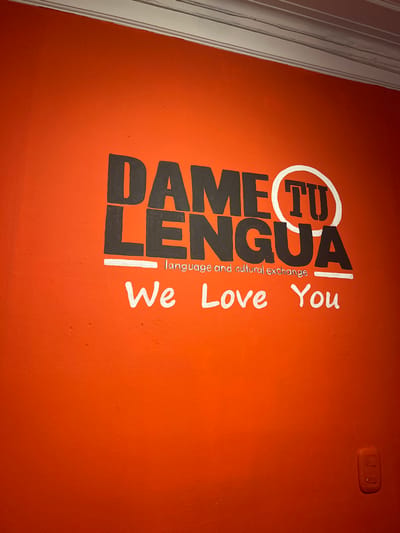The image displays an interior wall painted in a solid, vibrant orange hue, presenting a slightly textured finish. Near the top center, a logo and text are painted directly onto the wall. The primary text, "DAME TU LENGUA," is rendered in large, black, block-style letters with a worn or distressed appearance. The letters "TU" within "LENGUA" are encircled by a thick white outline. Below this, a smaller line of text in black, sans-serif font reads "language and cultural exchange." Further below, a third line of text in white, handwritten-style script states "We Love You."

Along the top edge of the wall, a white architectural crown molding or trim is visible. In the lower right quadrant of the image, a rectangular white electrical switch or outlet panel is affixed to the wall. No individuals are present or depicted, and no actions or interactions are occurring within the frame. The scene represents an interior space of the "Dame Tu Lengua" language and cultural exchange center, located in the city of Bogota, Capital District, Colombia.