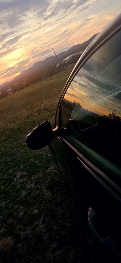 A vertical, low-angle shot captures a car's exterior against a sunset over a rural landscape. The car, predominantly black, is seen from its side, with the passenger door and side mirror visible. The side mirror is angled forward, reflecting the sky and the surrounding scenery. The car's window also mirrors the sunset, displaying warm hues of orange and yellow mixed with streaks of gray clouds.
Outside the car, a grassy field with patches of dirt stretches towards rolling hills in the distance. The hills are dotted with trees and small buildings, suggesting a village or town. A solitary utility pole stands against the sky on one of the hills. The sky itself is a dramatic canvas of a setting sun, casting a warm glow across the clouds and the landscape. The light is fading, creating a contrast between the illuminated sky and the shadowed foreground dominated by the car. The image appears to be taken in the vicinity of Momin Prohod, Bulgaria, given the landscape's characteristics. There are no people visible in the image.