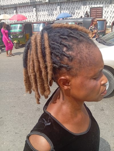 The image depicts an outdoor urban street scene in Ura Nta Umuarandu, Nigeria, under daylight conditions. The foreground features the right profile of a dark-skinned individual, whose head and right shoulder occupy the right half of the frame. This individual has two-toned dreadlocks (black and light brown/blonde) pulled back, and wears a black garment with a wide neckline and cut-out shoulders. The right ear is visible, and the eyes appear to be closed or downcast.
In the midground, an asphalt road surface is visible with several vehicles and pedestrians. Two light green/yellow auto-rickshaws (Keke Napep) are positioned on the left, one displaying "27" on its windshield. A darker auto-rickshaw is further back. A light-colored car, possibly beige or white, is partially visible on the far right, behind the foreground individual, showing a portion of its tire.
Multiple pedestrians are present. On the left, a woman in a pink form-fitting dress and sandals walks towards the right. Another woman, wearing a patterned dress and glasses, walks towards the left further to the right. Other indistinct figures are visible throughout the background.
The background consists of a building with a grey and white geometric patterned wall. Three umbrellas, colored red, blue, and light beige, are visible, suggesting shade structures. A brown door or gate is discernible on the right side of the building.
