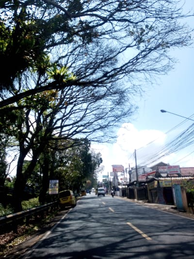 The image shows a road in Zeyathiri District, Myanmar, lined with buildings on the right and trees on the left. A yellow car is parked on the left side of the road. A white SUV and a motorcycle are traveling down the road. Overhead power lines run along the right side of the street. An "Alfamart" sign is visible. The sky is partially cloudy. The road is asphalt with a yellow dividing line. The scene is in daylight.