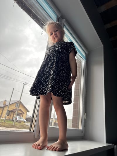 A young, blonde-haired child is positioned on a white windowsill, looking into the camera. She is wearing a dark, spotted dress. Outside the window, a view of a yellow brick house and a white car are visible. The location is likely Gornyi (Dvurechenskoe S/P), Russia, based on the prompt. The sky is overcast.