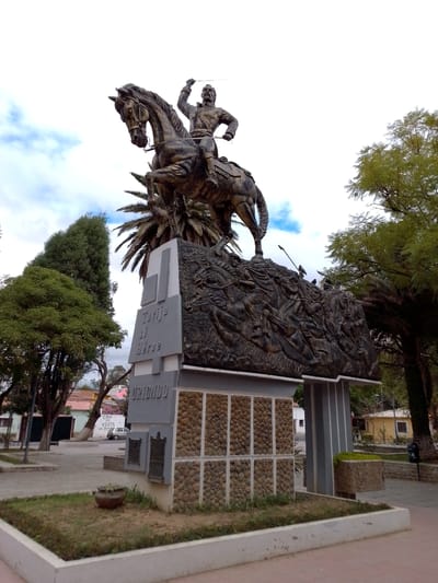 A statue of a man on horseback stands atop a monument in what appears to be a town square or park in Municipio Tarija, Bolivia. The statue itself is bronze-colored and depicts a man in military attire with his right arm raised, brandishing what appears to be a sword. The horse is rearing up, adding to the dynamic pose of the statue.

The monument beneath the statue is constructed of stone and concrete. It features relief carvings of people, possibly depicting a battle or historical event. The monument includes the words "TARIJA AL HEROE" and "URIONDO" in raised letters. The base of the monument is decorated with smaller stones arranged in a grid pattern.

The surrounding environment includes trees, plants, and a paved area that looks like a town square. In the background, there are buildings and a car, indicating that this location is within a populated area of Municipio Tarija. The sky is partly cloudy.