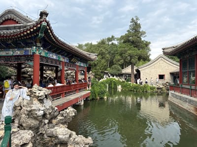 This image depicts a scenic view of a Chinese garden in Xicheng District, China, likely the Beihai Park, known for its imperial garden features. In the foreground, a woman in a traditional light blue dress stands near a large, irregularly shaped rock formation. She is holding a fan and appears to be posing. Behind her, a red-railed walkway is visible, with several people strolling along it. The walkway overlooks a body of calm, green water.
To the left, a traditional Chinese pavilion with ornate red pillars, intricate colorful carvings, and a tiered, tiled roof dominates the scene. This structure is supported by the rocky landscape. In the background, more traditional Chinese architecture can be seen, including buildings with grey tiled roofs and light-colored brick walls, partially obscured by lush green trees. The overall environment suggests a serene and picturesque garden setting. The sky is overcast with clouds, indicating a diffused light. The image captures a moment of cultural appreciation and leisurely activity within a historical Chinese garden.