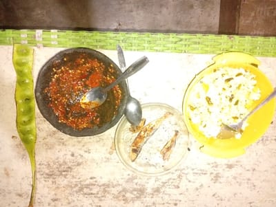 A top-down shot reveals a spread of Indonesian cuisine. A long, green petai bean rests to the left of a dark stone bowl filled with vibrant red sambal and a spoon. A clear glass plate holds three small, fried fish. To the right, a yellow bowl contains rice mixed with green vegetables, also with a spoon. The food is arranged on a weathered white surface, likely a table.