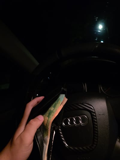 The image captures a close-up, low-light scene inside a car, primarily focusing on a human hand holding an open black leather wallet. The wallet contains several banknotes, predominantly green ones consistent with Euro currency, likely 20 Euro notes, though specific denominations are not fully legible.
The hand, with light to medium skin tone, is positioned in the lower right portion of the frame. To the left and upper part, the interior of a car is visible. A black steering wheel with a textured surface dominates this area, featuring the distinctive silver Audi four-ring logo on its airbag cover. The logo is framed by a decorative carbon fiber pattern trim. Partial text "AIRBAG" is visible on the steering wheel, along with a small, stylized horn symbol.
The overall environment is very dark, suggesting it is nighttime or a poorly lit interior. Bright, out-of-focus lights are visible outside the car on the far left, reflecting off what might be a window. The scene depicts a simple, everyday activity of handling money inside a vehicle. The presence of Euro banknotes, in the context of Sevlievo, Bulgaria, suggests common usage of the currency in the region. No explicit emotions are discernible.