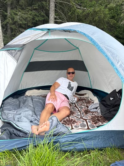 A man in a white t-shirt and pink shorts is lying inside a tent. He is wearing sunglasses and has one leg crossed over the other. There is a sleeping bag and a blanket on the floor of the tent. A black backpack is next to him. The tent is set up in a grassy area with trees in the background, suggesting a natural, outdoor setting, possibly near Sarnitsa, Bulgaria, given the context of the prompt.