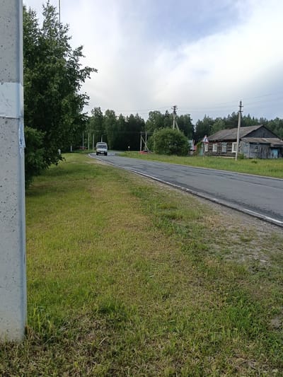 A roadside scene in Красный Маяк, Russia, shows an asphalt road curving slightly to the left with grassy verges on either side. A light-colored van approaches on the road. To the right of the road is a weathered wooden building with visible windows and what appears to be an extension. Utility poles line the road. The sky is overcast. A concrete pole stands in the foreground to the left. A dense line of trees appears in the background. The overall impression is of a rural or suburban setting.