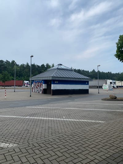 The image shows a small, octagonal building with a multi-pitched roof and a glass ceiling, situated in an outdoor paved area. The building has a mural on its side, with blue, white, and black stripes. To the left of the building are two lampposts. In the background, there are trees, a long red shipping container on a truck, and several white tractor-trailer trucks. The paved area is made of interlocking concrete blocks and has white lines painted on it, indicating parking spaces. A large grey rock is visible in the foreground to the right. The sky is overcast. The building appears to be a public restroom facility, likely located at a rest stop or service area, possibly on a highway. The graffiti on the building suggests it is in an urban or semi-urban area. The presence of trucks and shipping containers suggests it is a transportation hub or a facility used by commercial vehicles. The environment is clean and well-maintained, with no visible litter or damage. The overall impression is one of a functional and utilitarian space.