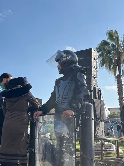 An outdoor scene in Al-Qanawat Municipality, Syria, features a male police officer in riot gear standing in the foreground, facing right in profile. He wears a black helmet with "POLICE" in white, a raised clear visor, and black protective padding on his torso, shoulders, and arms. He has a beard and holds a transparent riot shield with both hands, resting it on a dark metal pole. Behind him to the right, a tall stack of black sound speakers is visible, next to a palm tree. To the left of the officer, the back of a person in a brown fur-lined hooded jacket is partially visible, along with another individual's head. The ground is grassy. In the background, several indistinct people are present on a field, and a light-colored building is partially visible under a clear blue sky with minimal clouds. Metal chains connect dark poles, forming a barrier in the lower mid-ground.