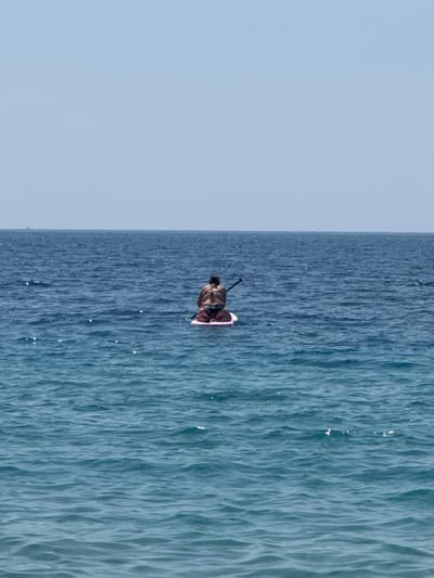 A person is paddleboarding on the sea. The person is sitting on the paddleboard, facing away from the camera. They are wearing a swimsuit. The paddleboard is pink and white. The sea is blue and calm. The sky is clear and blue. The image is taken from a distance, so the person's features are not visible. There is a distant object visible on the horizon in the far distance to the left.