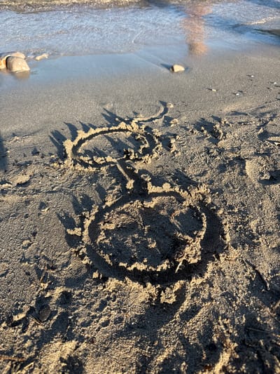 This image depicts a beach scene in Donji Katun, Montenegro, under natural daylight conditions, likely morning or late afternoon given the elongated shadows. The foreground consists of light brown-grey sand, textured with visible grains and small debris. Two figures are drawn into the sand: a large, roughly circular shape, approximately 30-40 cm in diameter, and a smaller, less defined loop positioned above and slightly to the left, connected by a narrow line or stem. These forms create distinct, dark shadows stretching towards the lower right of the frame. Towards the midground, the sand transitions to a darker, wetter texture as it meets the water's edge. Several small stones and pebbles are visible in this area, including one larger, light-colored stone partially submerged in shallow water on the left side. The background features calm, light blue-grey water with subtle ripples, reflecting ambient light. No individuals or animals are visible.