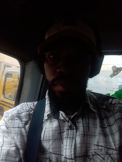 The image is a close-up, selfie-style shot of a man sitting in the driver's seat of a vehicle, likely a truck or bus, in Ikeja, Nigeria. The man is wearing a plaid shirt, a baseball cap, and headphones. He has a beard and mustache. The interior of the vehicle is dark, with some light coming from the outside. Through the windshield, another yellow vehicle is visible. There is also a window behind the man with a sticker on it. The scene appears to be a street in Ikeja, given the presence of vehicles and the context.