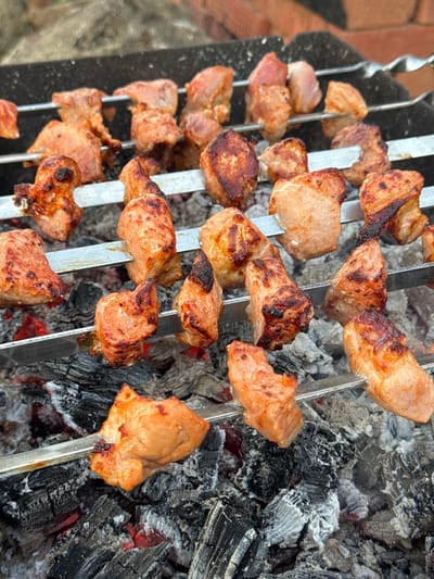 The image depicts a close-up, eye-level view of several skewers of meat being grilled over hot coals. The meat pieces are cubed and appear to be chicken or pork, with some pieces showing char marks from the grill. The skewers are arranged horizontally on a grill grate, with embers glowing beneath them. The background is slightly blurred, showing what appears to be a brick surface and some greenery, suggesting an outdoor setting. The overall impression is one of a barbecue in progress. No specific location in Maksimovo, Russia, is identifiable from the image alone.