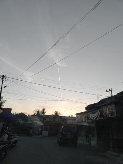 In the city of Kumbang, Indonesia, a paved road curves slightly through a sparsely populated urban area. The sky above is a muted gray with a faint hint of sunset orange peeking through from the horizon. A distinct contrail from an aircraft stretches vertically across the sky, a stark white line against the pale backdrop. Overhead, a complex network of black electrical wires crisscrosses the frame, supported by utility poles.

Along the right side of the road, a dimly lit building with a dark facade has a sign displaying text and the price "18.000". To the left, several motorcycles are parked on the side of the road. One of these is a white scooter. Further down the road, a dark-colored car is visible. In the distance, a person is riding a bicycle. The scene appears to be a street view, possibly captured from a vehicle, showcasing a slice of everyday life in this Indonesian locale. The overall atmosphere is subdued, with the fading light and the stark lines of the utility wires creating a somewhat industrial or functional aesthetic.