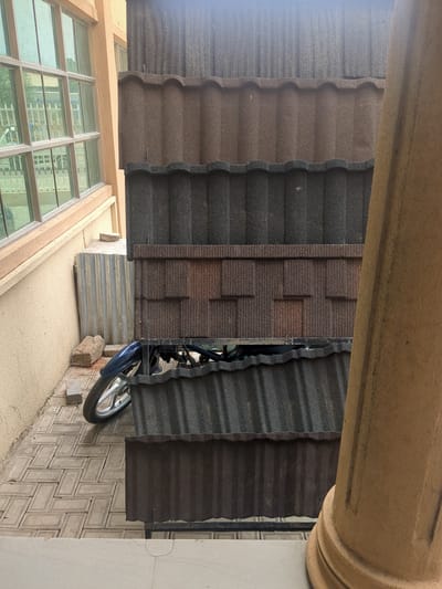 The image shows a collection of stone-coated roofing sheets displayed in a row. They are a mix of dark gray, brown, and a patterned brown variety. A motorcycle, partially visible, is parked behind some of the roofing sheets, with its front wheel and a portion of its blue bodywork in view. The scene appears to be an outdoor display, possibly at a building materials yard or a construction site, given the stacked roofing sheets and the presence of building elements like a window with reflective glass and a textured wall. The lighting suggests it is daytime, but the weather is not discernible. The image is oriented sideways.