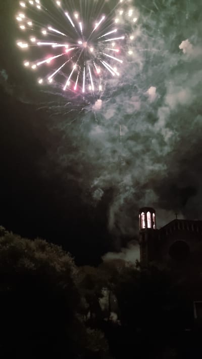 This media file captures a nighttime scene featuring a fireworks display. The foreground is dominated by a vibrant burst of fireworks, radiating bright white and pink lights against a dark sky filled with smoke and wisps of clouds. To the right of the frame, a silhouette of a building with an illuminated bell tower can be seen, suggesting a church or similar structure. Trees, also in silhouette, are visible in the lower right corner. The overall atmosphere is festive and celebratory, indicative of a special event. The time of day is clearly night, and the weather appears clear enough for fireworks. The location context provided, Sant Joan Les Fonts, Spain, points to a specific geographical setting for this event, potentially a local festival or holiday celebration. No text is visible in the image.