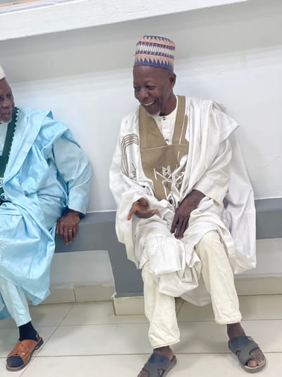 Two elderly men are depicted seated indoors in Zaria, Nigeria. The man on the right, with a dark complexion, wears a white traditional tunic featuring prominent brown embroidery, beige trousers, and a multi-colored geometric patterned cap. He is smiling broadly, exposing his teeth, with his left hand slightly open and extended forward, and his right hand resting on his lap. He wears dark slide sandals. The man on the left, partially visible, also dark-complexioned, wears a light blue traditional garment over a green undergarment and a white cap. A silver ring is visible on the ring finger of his left hand, which rests on his lap. He wears brown slide sandals over black socks. Both men are positioned against a plain, off-white wall with a horizontal relief detail and a grey lower section, above a light-colored tiled floor.