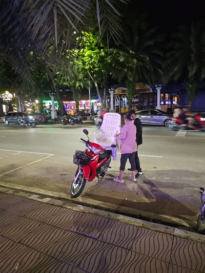 This media file captures a street scene at night in Ban Klong Haeng, Thailand. The image is oriented vertically, with the top of the frame showing palm fronds against a dark sky. In the mid-ground, two people are visible on the right side of a paved road. A person wearing a pink shirt and dark pants is standing behind a white plastic chair, facing away from the camera. Another person, in a dark jacket and dark pants, stands beside them, also facing away. A red motorcycle is parked on the right side of the road, in front of the people and the chair.

The background reveals a street lined with buildings, illuminated by various neon and colored lights, suggesting a commercial or entertainment district. There are other motorcycles and a silver car visible on the road, indicating traffic. The ground on the right is a patterned sidewalk. The overall atmosphere appears to be lively, with the artificial lighting creating a vibrant night scene. The time of day is clearly night, given the darkness of the sky and the extensive artificial lighting. The weather is not discernible from the image.