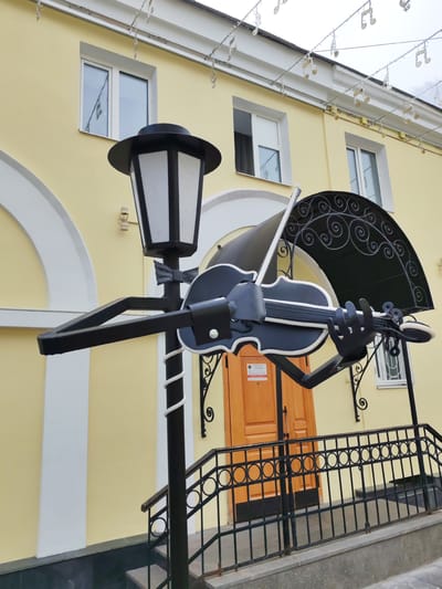 The image shows a street art installation in Yaroslavl, Russia. The sculpture depicts a lamppost with a metal violin and bow held by two robotic hands. The lamppost is decorated with a black bow tie and a white spiral stripe. In the background, a light yellow building with white accents and arched windows is visible. A wooden door and a black metal railing with circular patterns leading up stairs are also present. Above the building, strings with musical note decorations hang across the street. The overall scene suggests a creative and artistic ambiance in a public space.