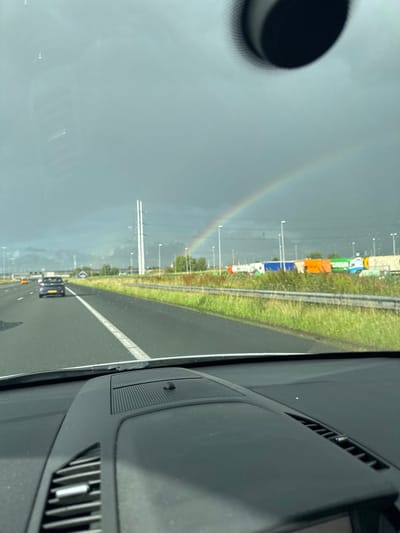 The image captures a view from inside a vehicle traveling on a highway. A faint rainbow is visible in the distance, arching across a dark, cloudy sky. Several vehicles, including cars and trucks, are on the highway, and there are power lines and tall structures on the side of the road. The foreground shows a portion of the car's dashboard. The scene appears to be the A4 highway near Leiderdorp, Netherlands, given the characteristic Dutch landscape and infrastructure, including the distinctive high-voltage pylons. The overall atmosphere is one of travel under overcast conditions with a touch of natural beauty from the rainbow.